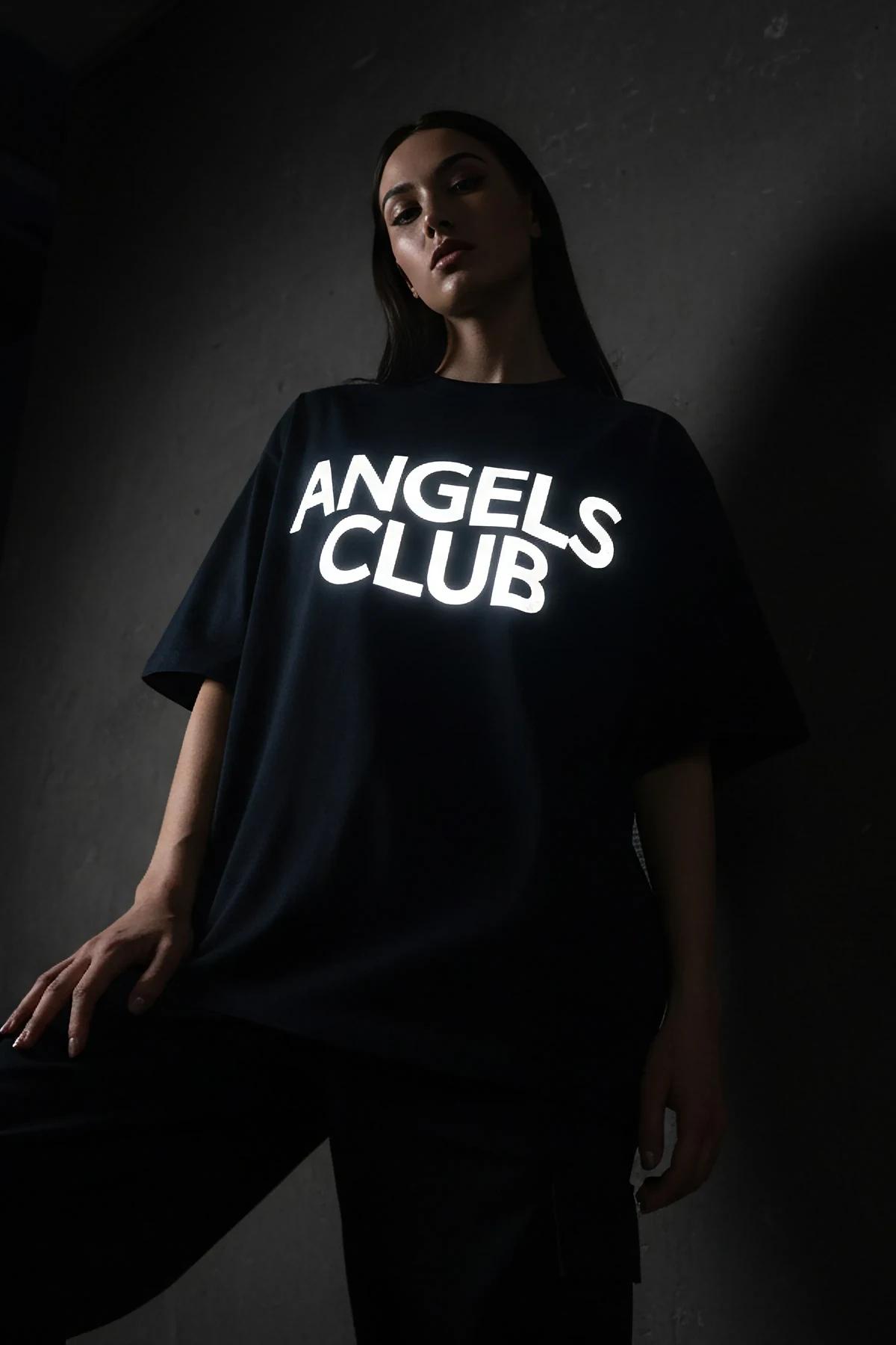 Işık altında parlayan siyah pamuklu oversize tişörtteki reflektör Angels Club göğüs baskısının yakın çekimi