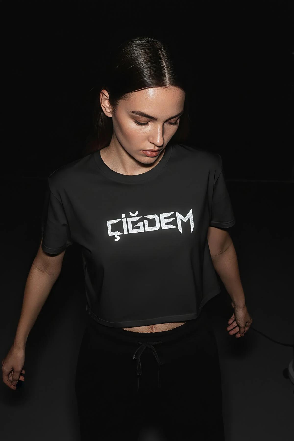 Kadın modelin üzerinde, tam göğüs genişliğinde kalın beyaz reflektör CIGDEM isim baskılı siyah pamuklu crop tişört
