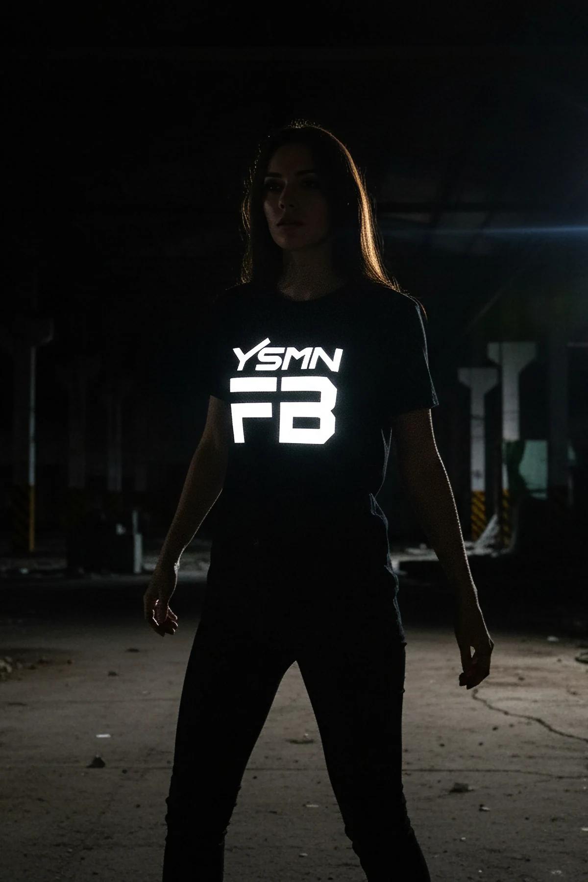 Karanlık ortamda kadın modelin üzerinde, doğrudan ışık altında beyaz parlayan kalın YSMN FB logo baskılı siyah reflektör grafik tişört