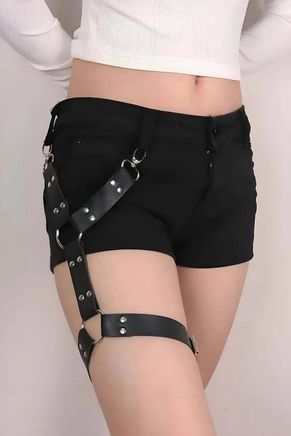 Siyah şort üzerinde gümüş O-halkalar ve perçinli zımbalarla siyah suni deri tek bacak harness, kalçadan uylukla ayarlanabilir bantlar