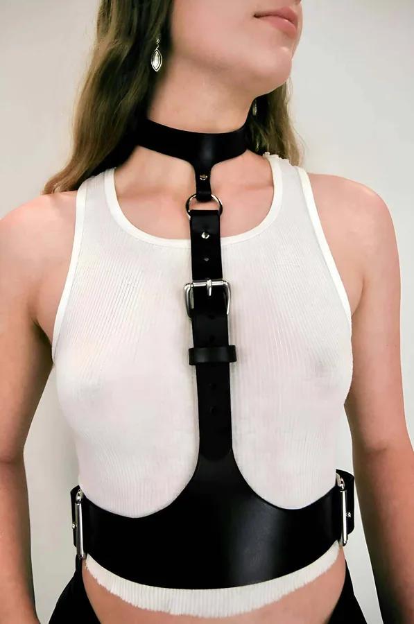 Beyaz üstün üzerine giyilmiş gümüş dikdörtgen toka, geniş bel bandı ve entegre çoker askılı siyah suni deri vücut harness'ı