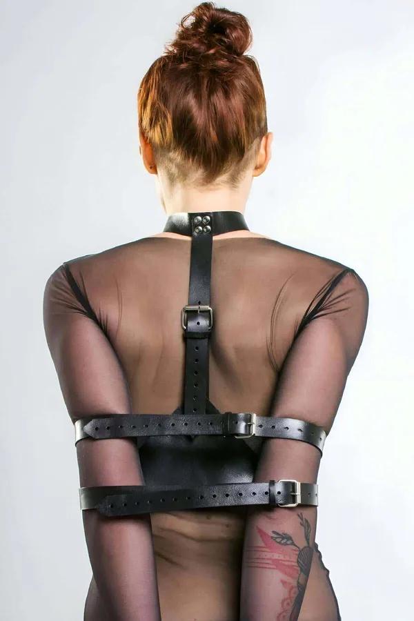 Şeffaf file üstün üzerine giyilmiş geniş kol kelepçeleri ve gümüş rengi tokalar ile siyah vegan deri arka harness, arka görünüm