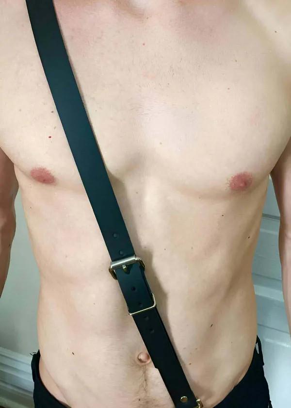 Erkek gövdesinde altın tonlu tokalı tek çapraz göğüs askılı siyah suni deri harness